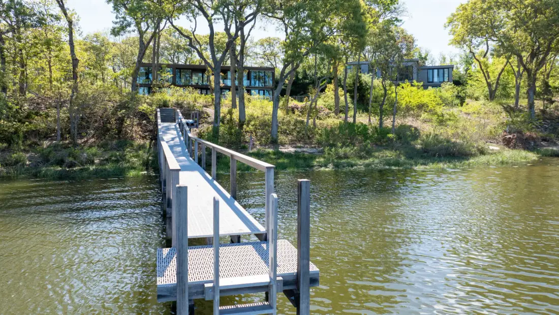 A narrow footbridge extends across a calm river toward a wooded bank.