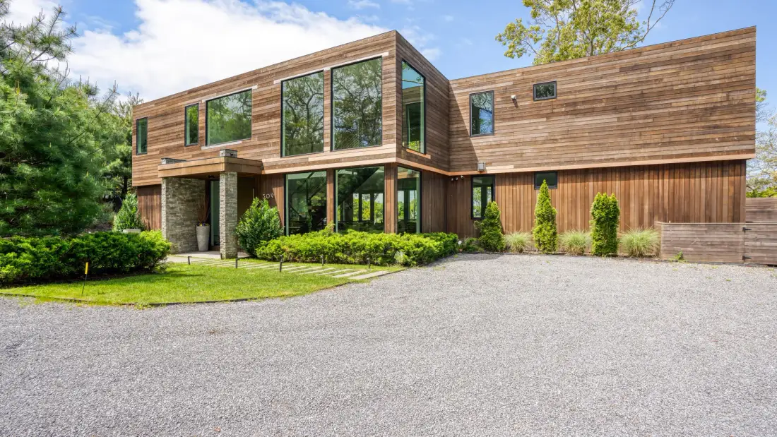 A modern wooden house with large windows faces a broad gravel driveway.