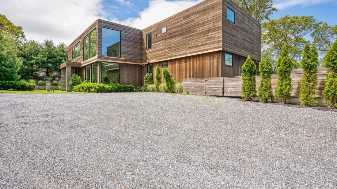 A modern wood-clad house stands beside a broad gravel driveway and landscaped greenery.