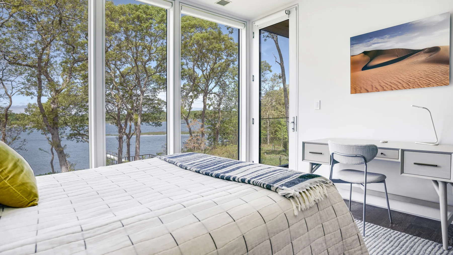 A bright bedroom has a bed beside large windows overlooking trees and water.