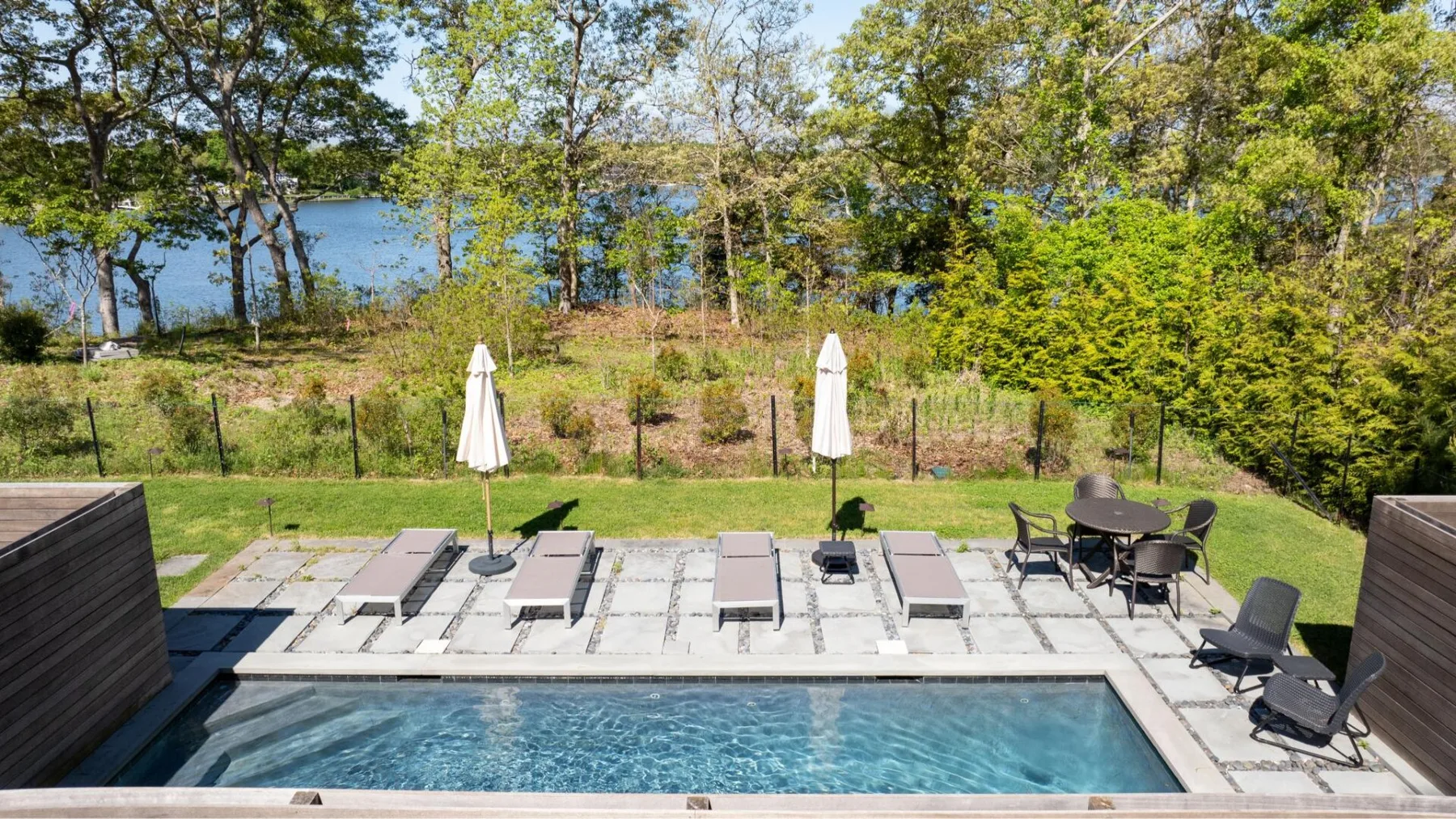 A swimming pool and paved patio with lounge seating overlook a tree-lined body of water.
