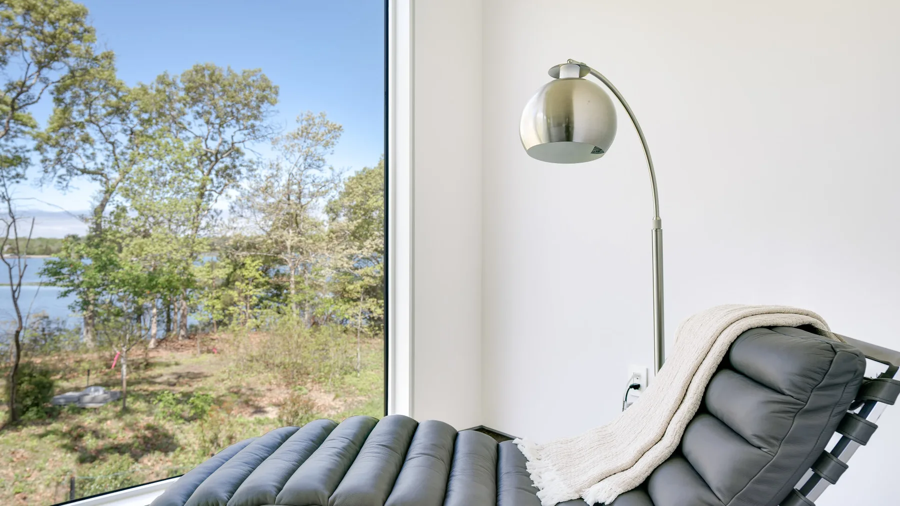 A cushioned lounge chair sits beside a large window overlooking trees and water.