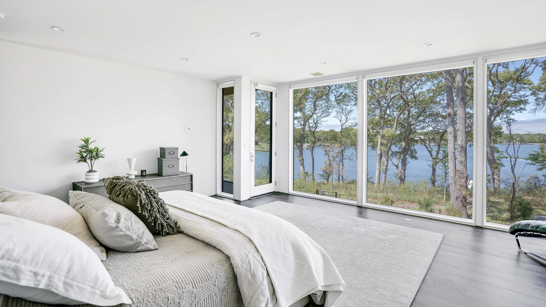 A bright bedroom with large windows overlooking trees and a lake.