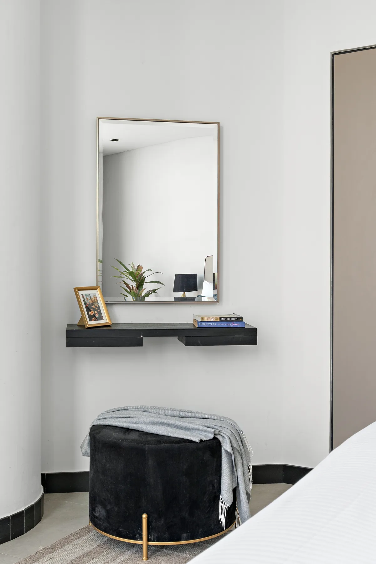 A bedroom corner with a round upholstered stool and a wall mirror above a narrow shelf.