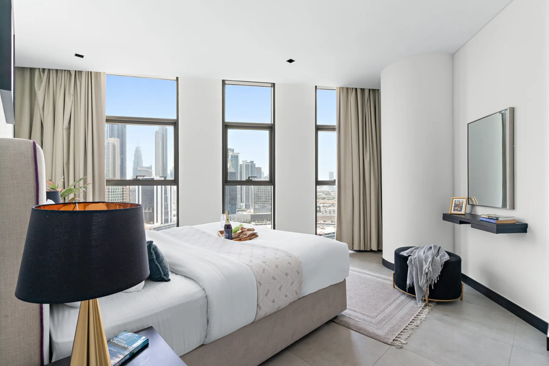 A bright bedroom with a neatly made bed and tall windows overlooking the city.