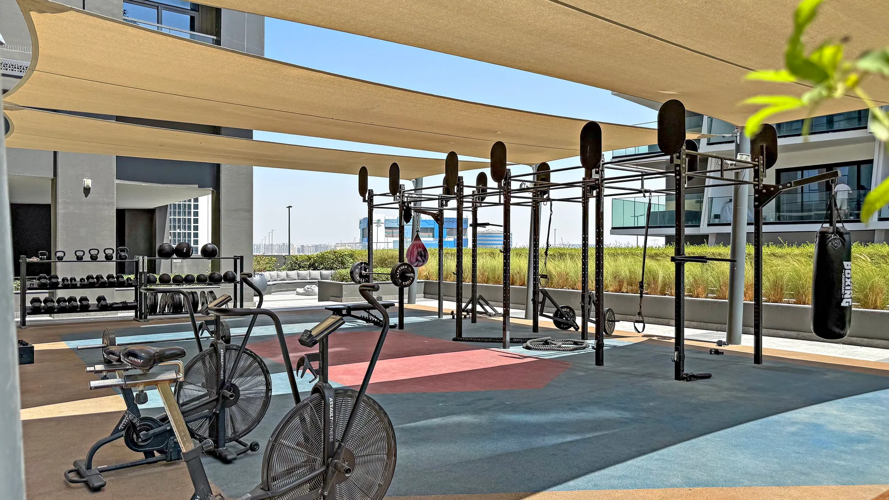 An outdoor fitness area with exercise bikes and a weight-training rig under shade sails.