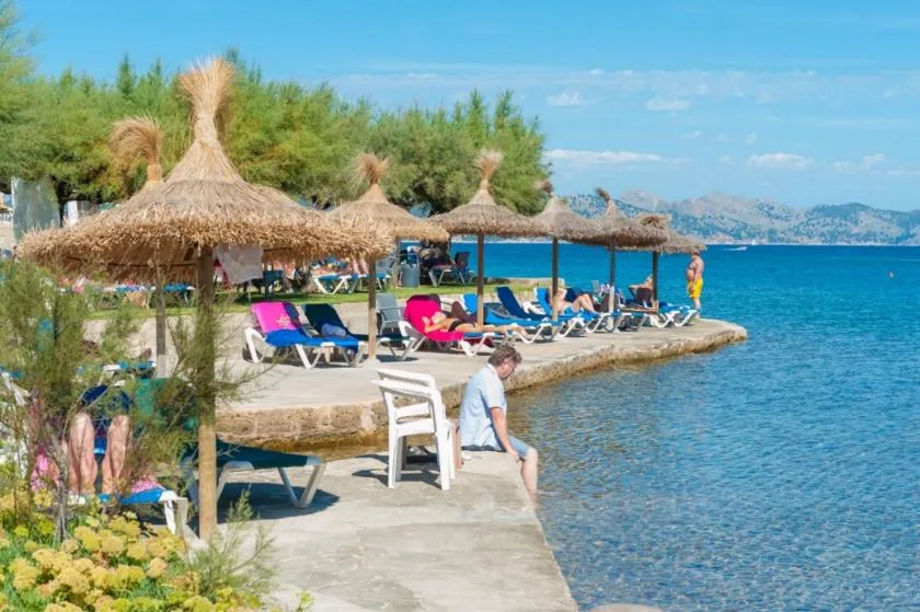 A seaside bathing area has lounge seating beneath thatched parasols beside clear blue water.