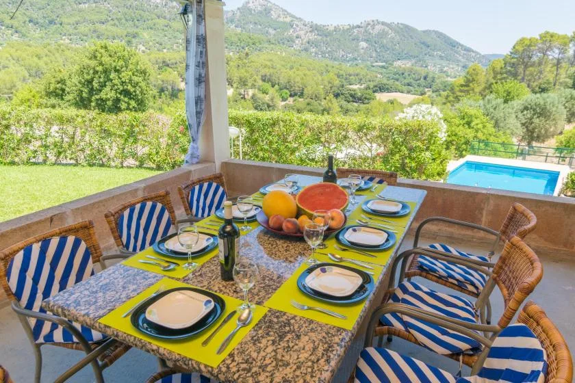 Comedor exterior cubierto con la mesa puesta, con vistas a un jardín y una piscina.