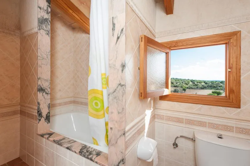Un baño alicatado con bañera, cortina de ducha y una ventana abierta con vistas a zonas verdes.