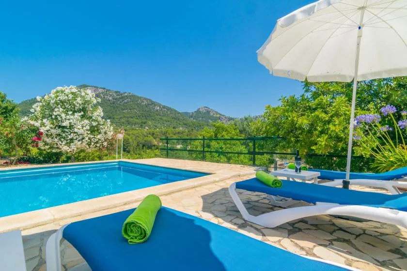 Una piscina al aire libre con tumbonas azules, una sombrilla y una colina verde al fondo.