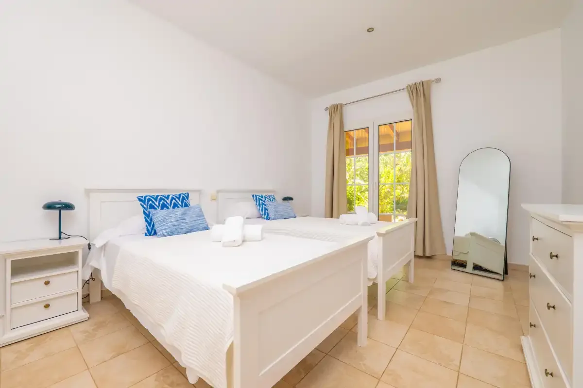 Un dormitorio luminoso con ropa de cama blanca y una ventana enmarcada por cortinas beige.