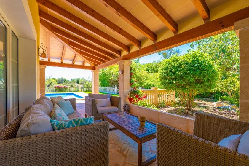 Un patio cubierto tiene asientos tejidos alrededor de una mesa baja, con vistas a un jardín y una piscina.