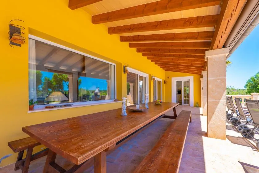 Un patio cubierto con una larga mesa de comedor de madera junto a paredes amarillas y ventanales.