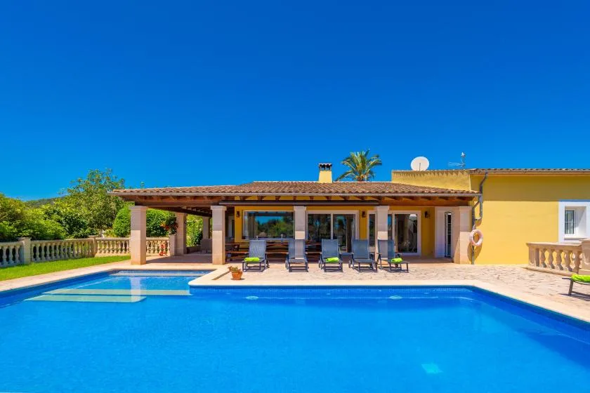 Una casa con patio cubierto está junto a una piscina bajo un cielo azul despejado.