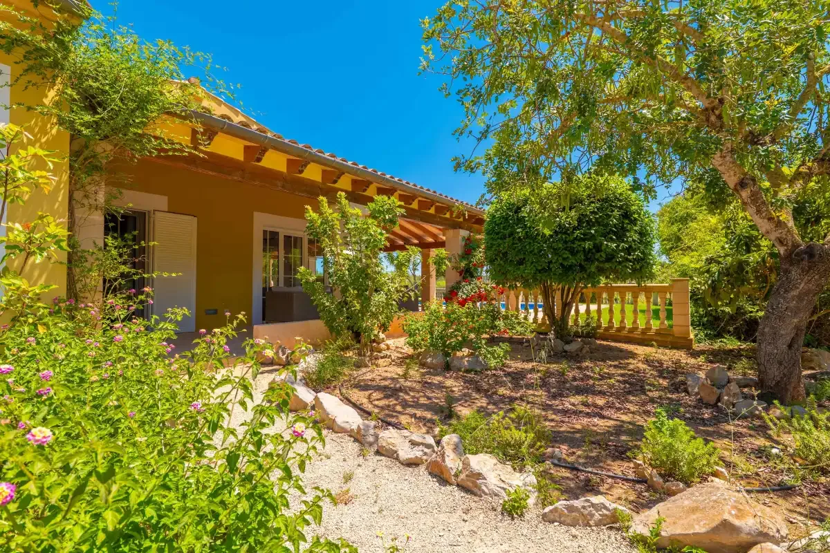 Un patio ajardinado junto a una casa amarilla, con plantas en flor, árboles y una barandilla de madera.