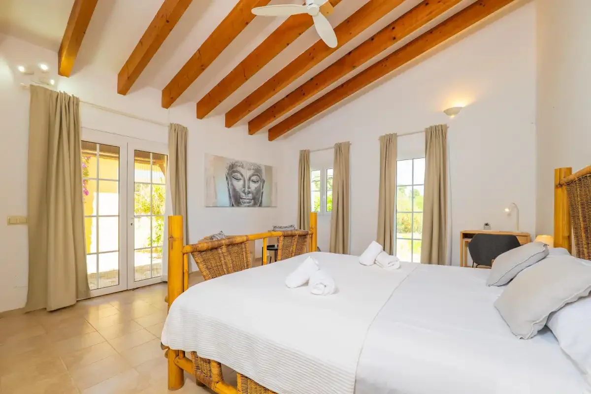 Un dormitorio luminoso con ropa de cama blanca, vigas de madera vistas y ventanas altas con cortinas.