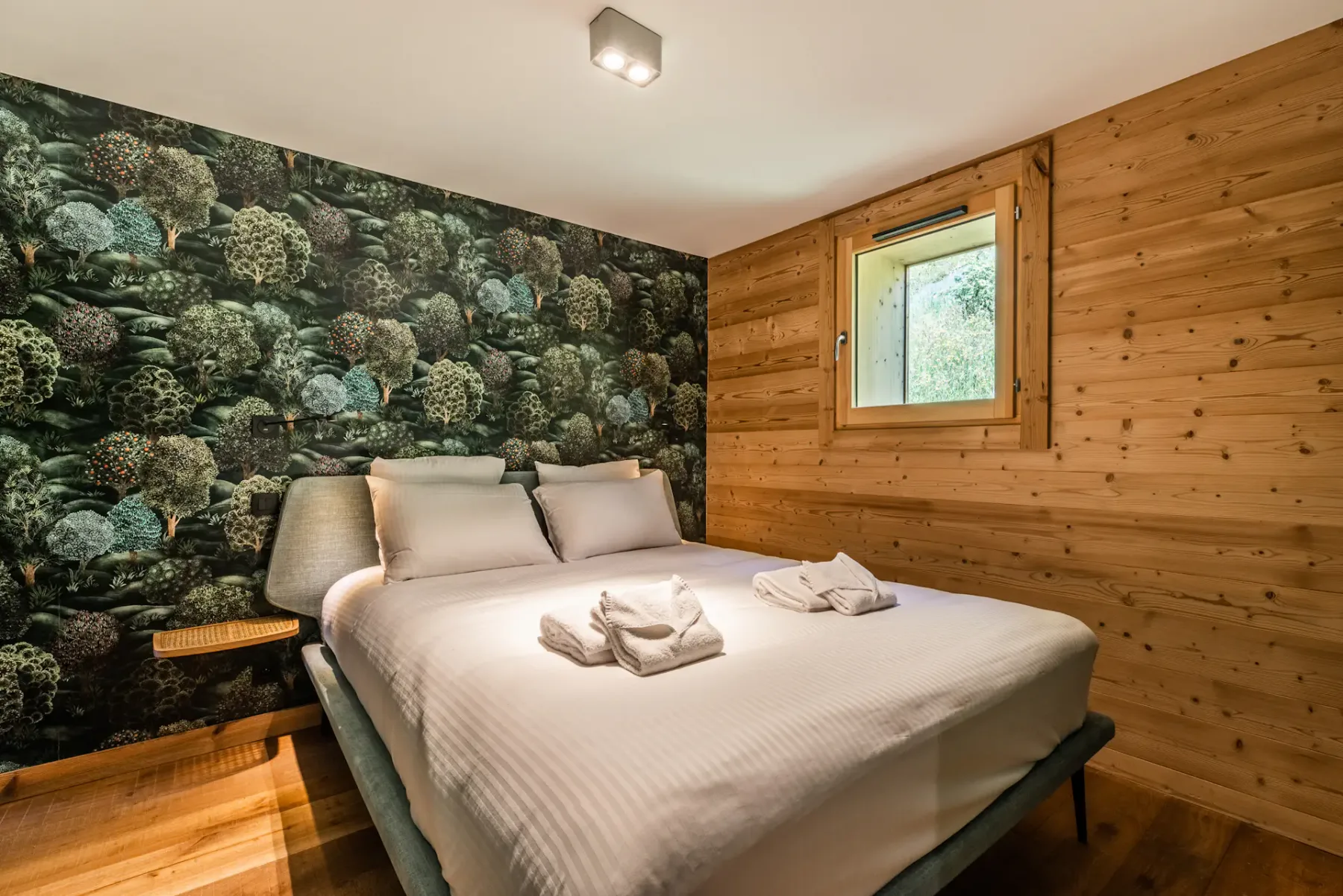 A bedroom with a neatly made bed and forest-patterned wallpaper.