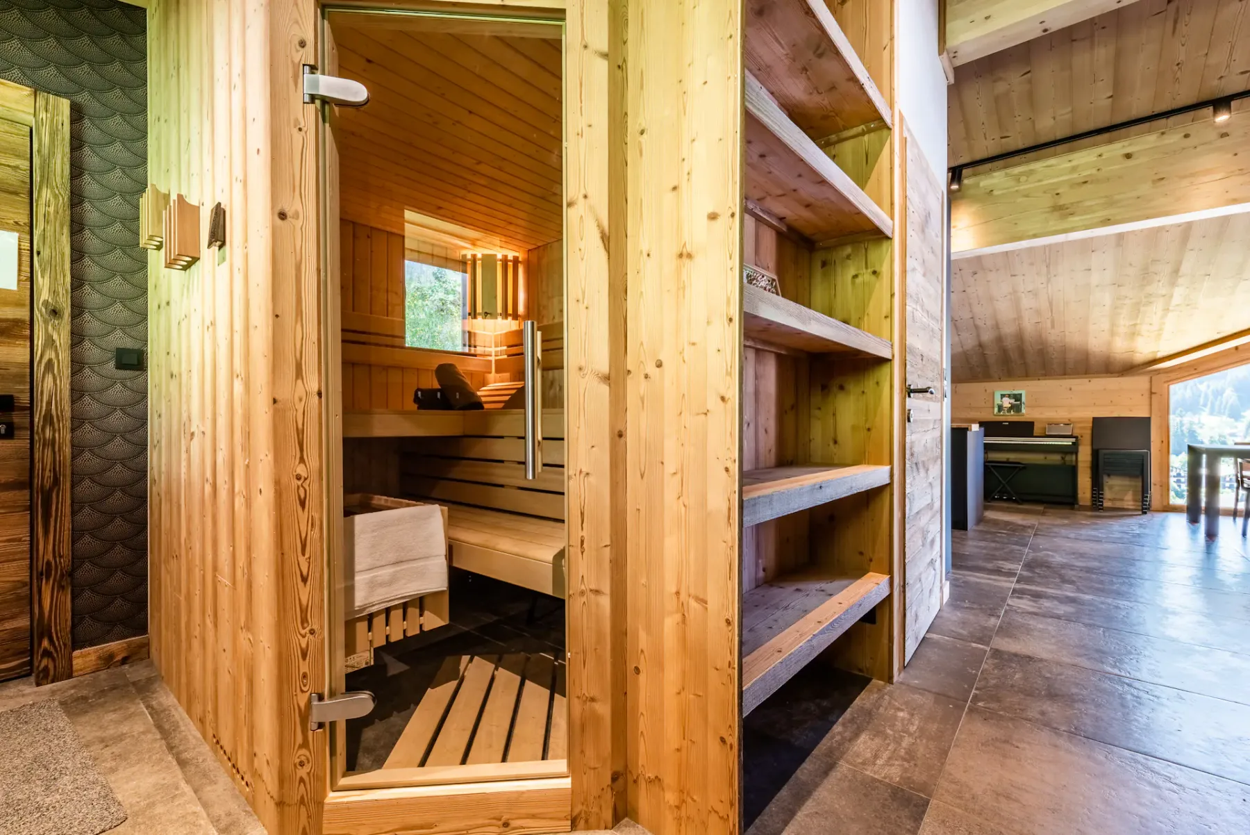A wooden sauna room with benches and a glass door opens onto a tiled interior.