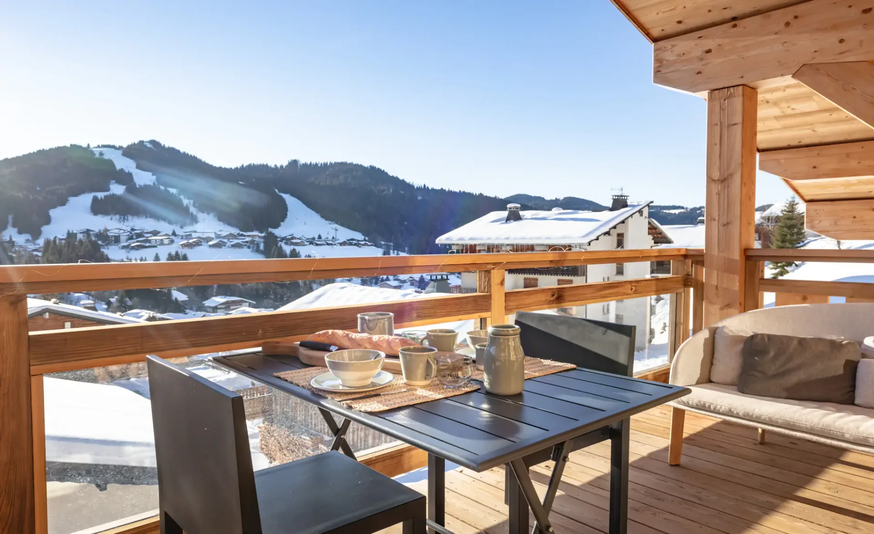 A wooden terrace with outdoor seating and a table overlooks snowy mountain slopes.