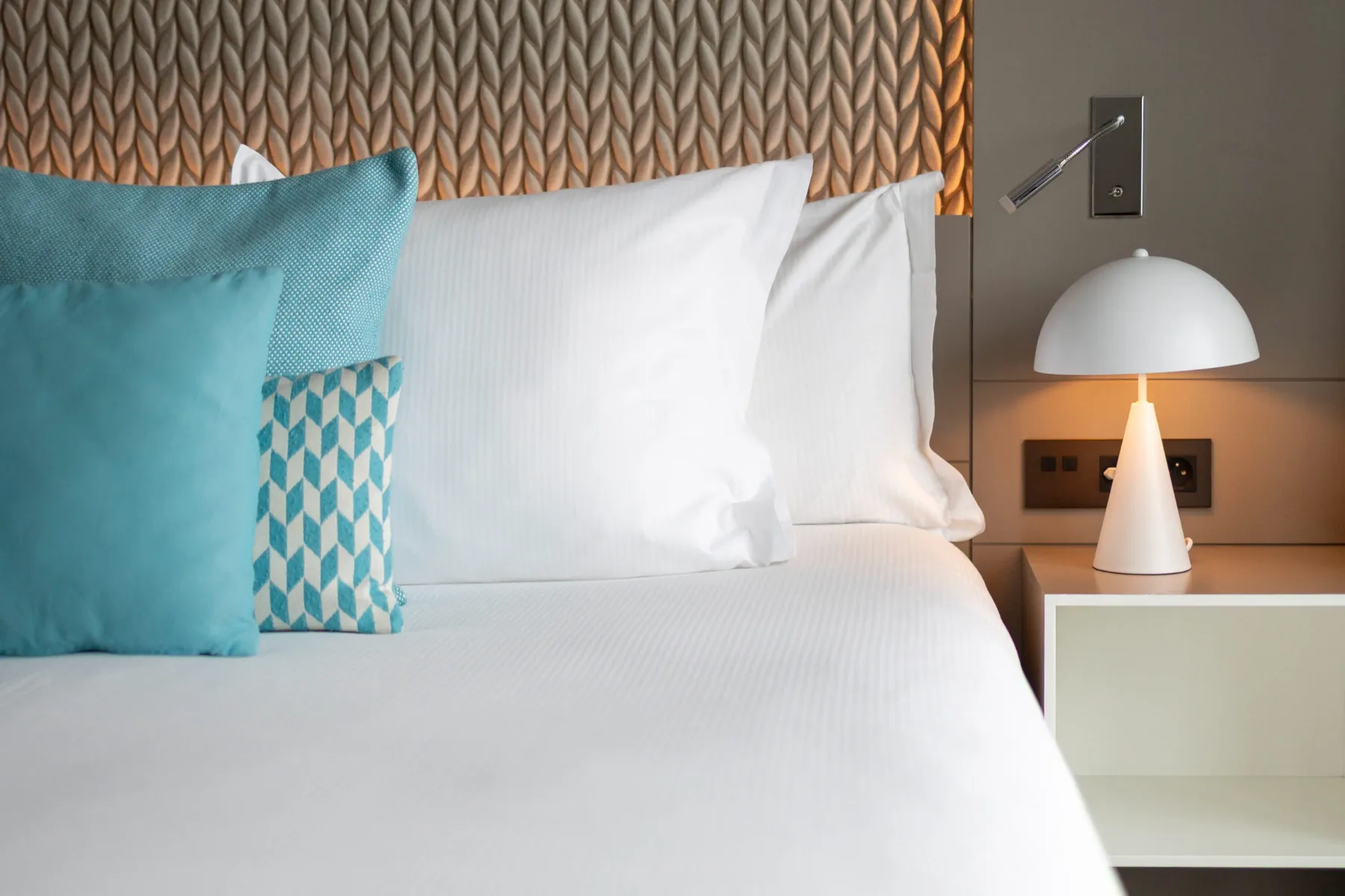 Cama de hotel con cabecero tapizado y lámpara de noche.
