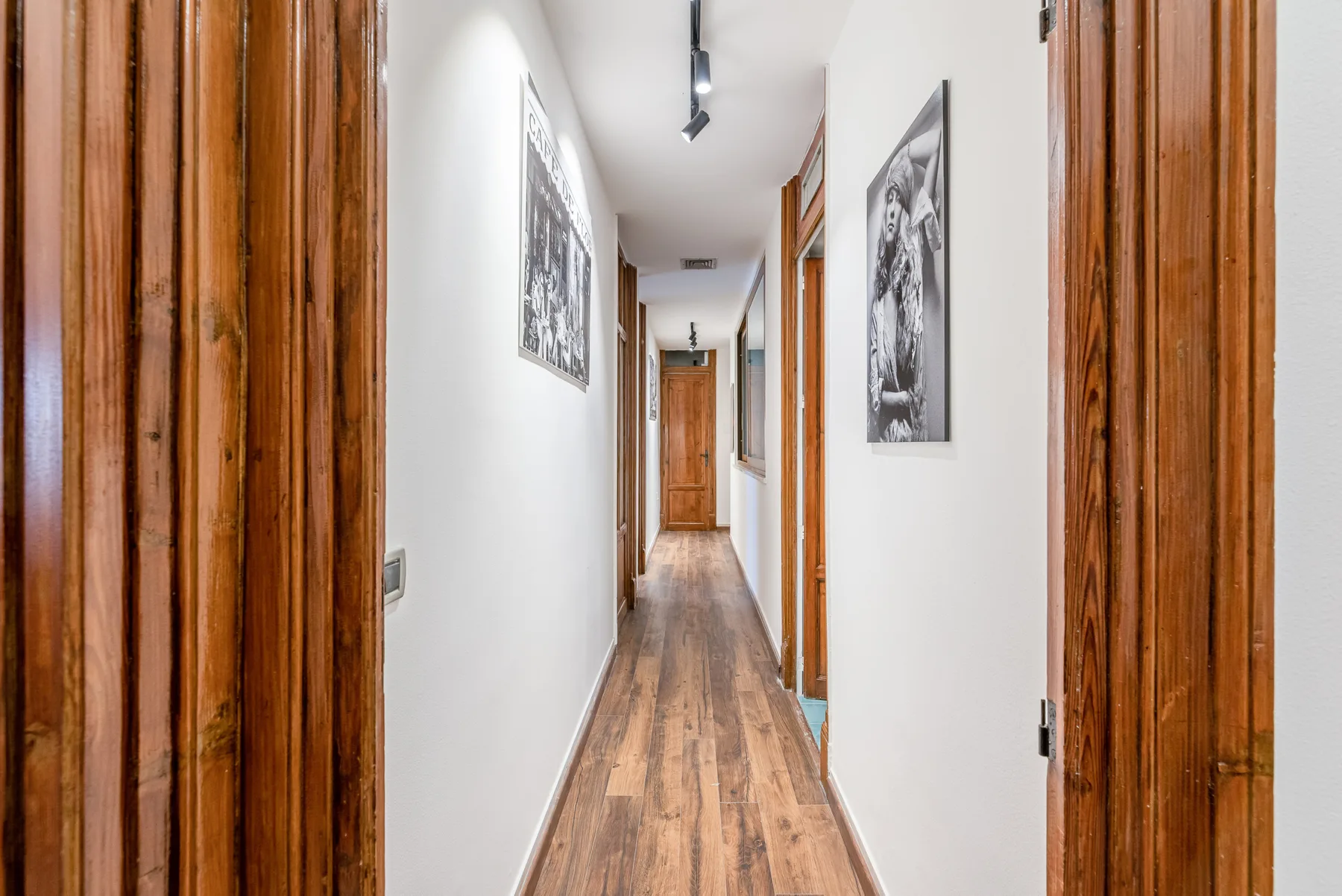 A narrow hallway with wood flooring, framed pictures, and several doorways.
