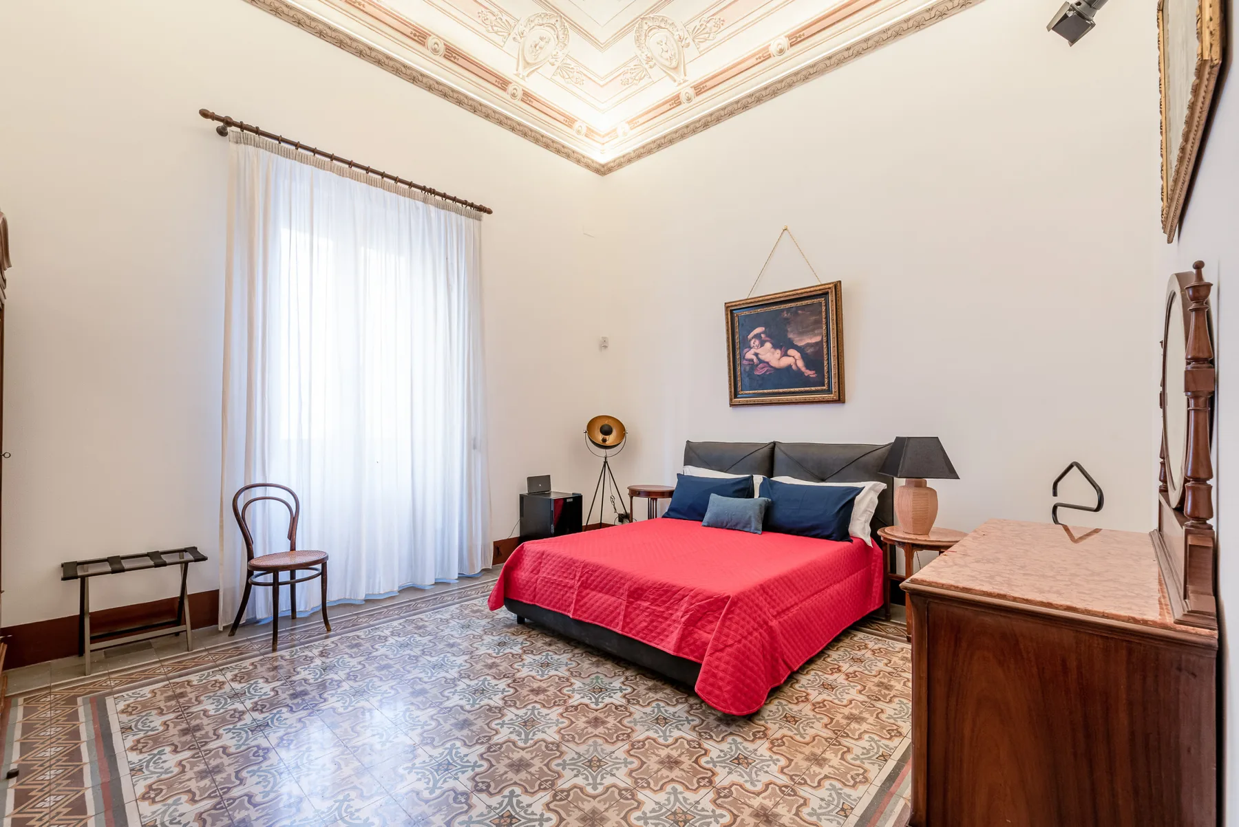 A bedroom with a bed covered in red bedding and a tall curtained window.