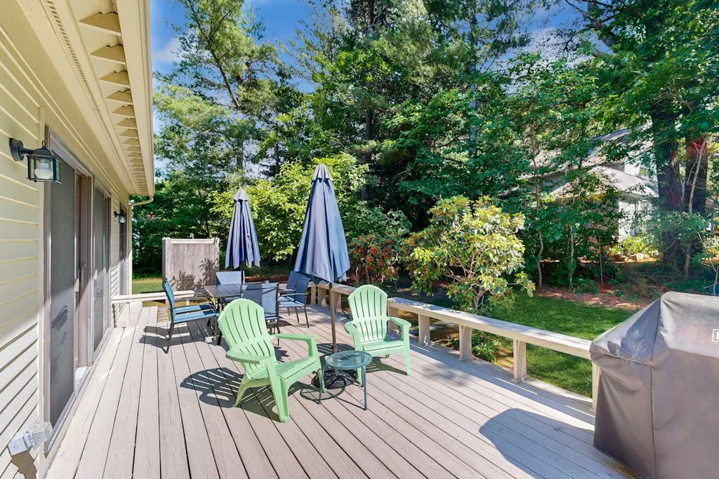 An outdoor deck with green seating and closed umbrellas overlooks a leafy yard.