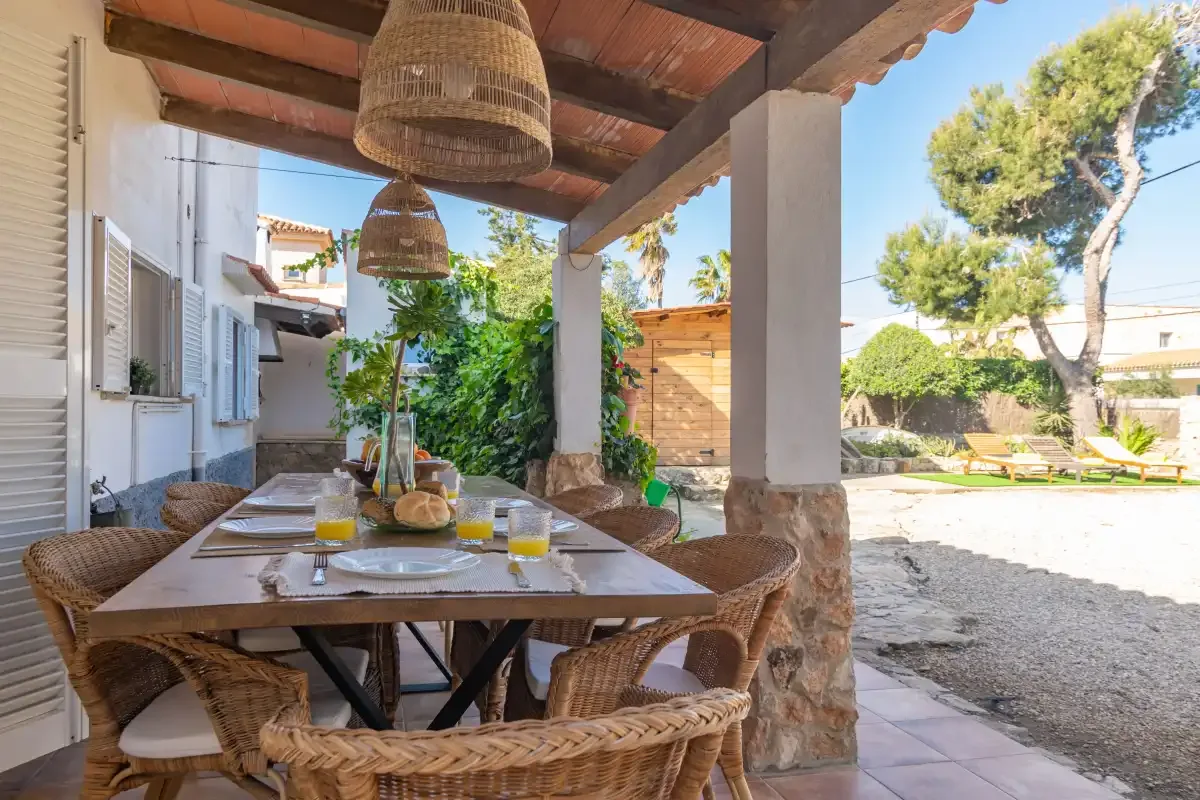 A covered outdoor dining area with a table and woven seating overlooks a sunny yard.