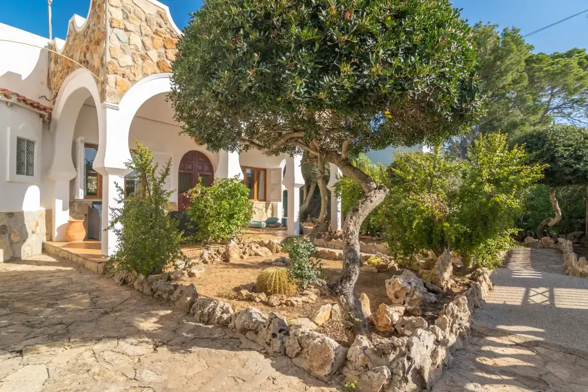 A white house entrance with arched openings sits beside a leafy tree and stone path.