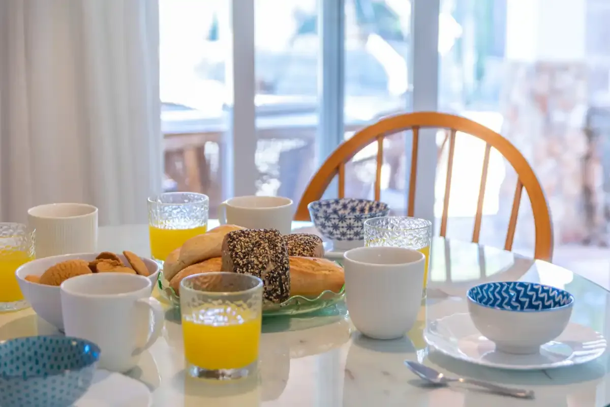 A dining table is set for breakfast with bread, juice, cups, and bowls.