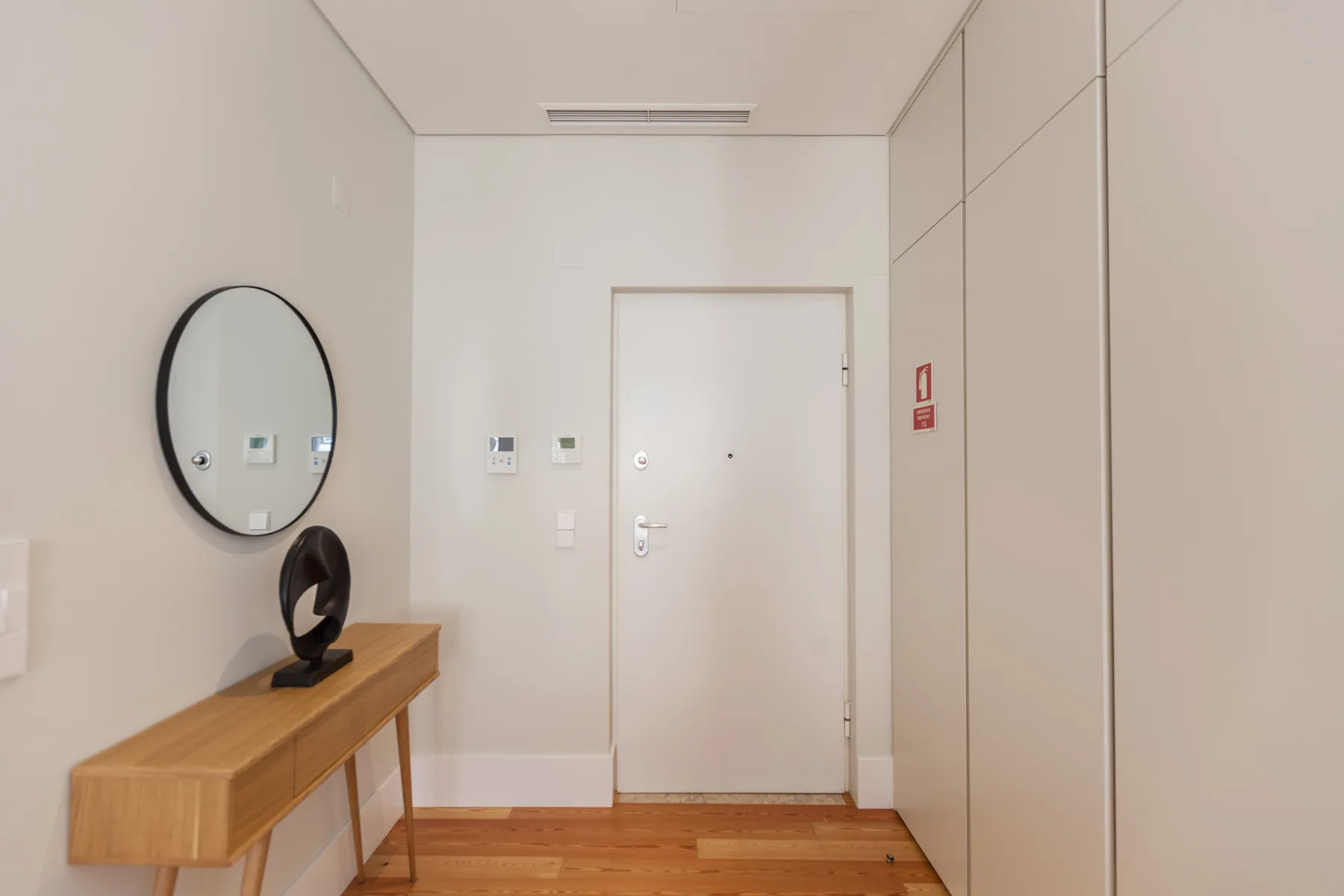 Una entrada de apartamento de tonos claros con una consola de madera y un espejo redondo.