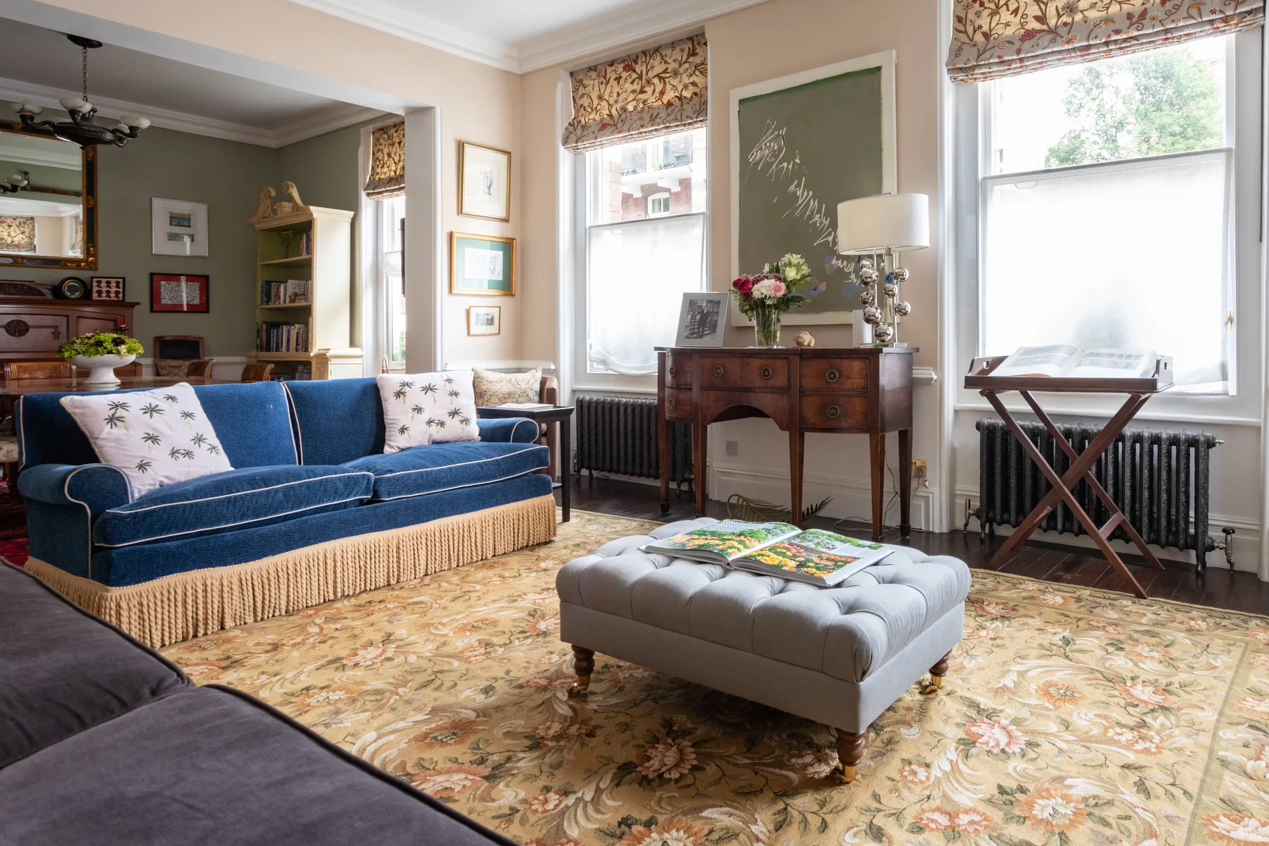 A living room with blue seating, a tufted ottoman, and tall windows.