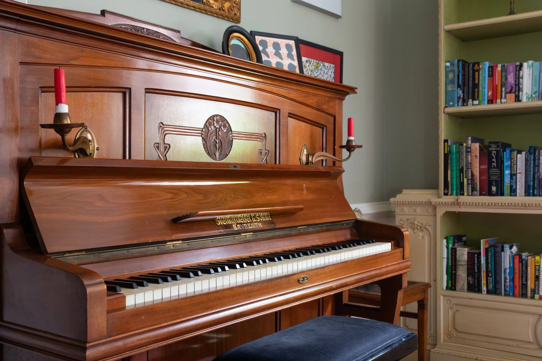 A wooden upright piano with a dark bench stands beside a bookshelf.