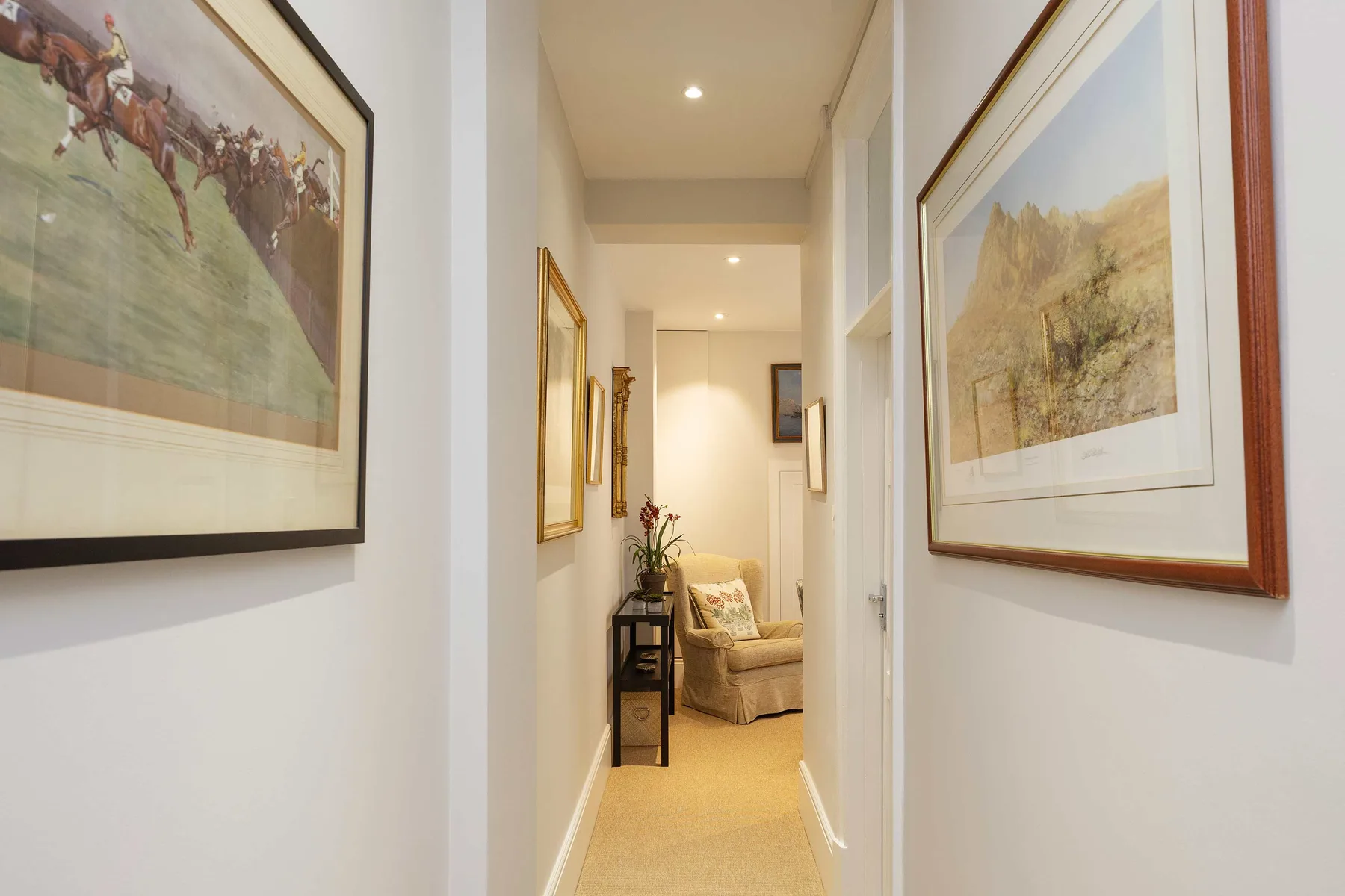 A narrow hallway with framed artwork leads to a small seating area.