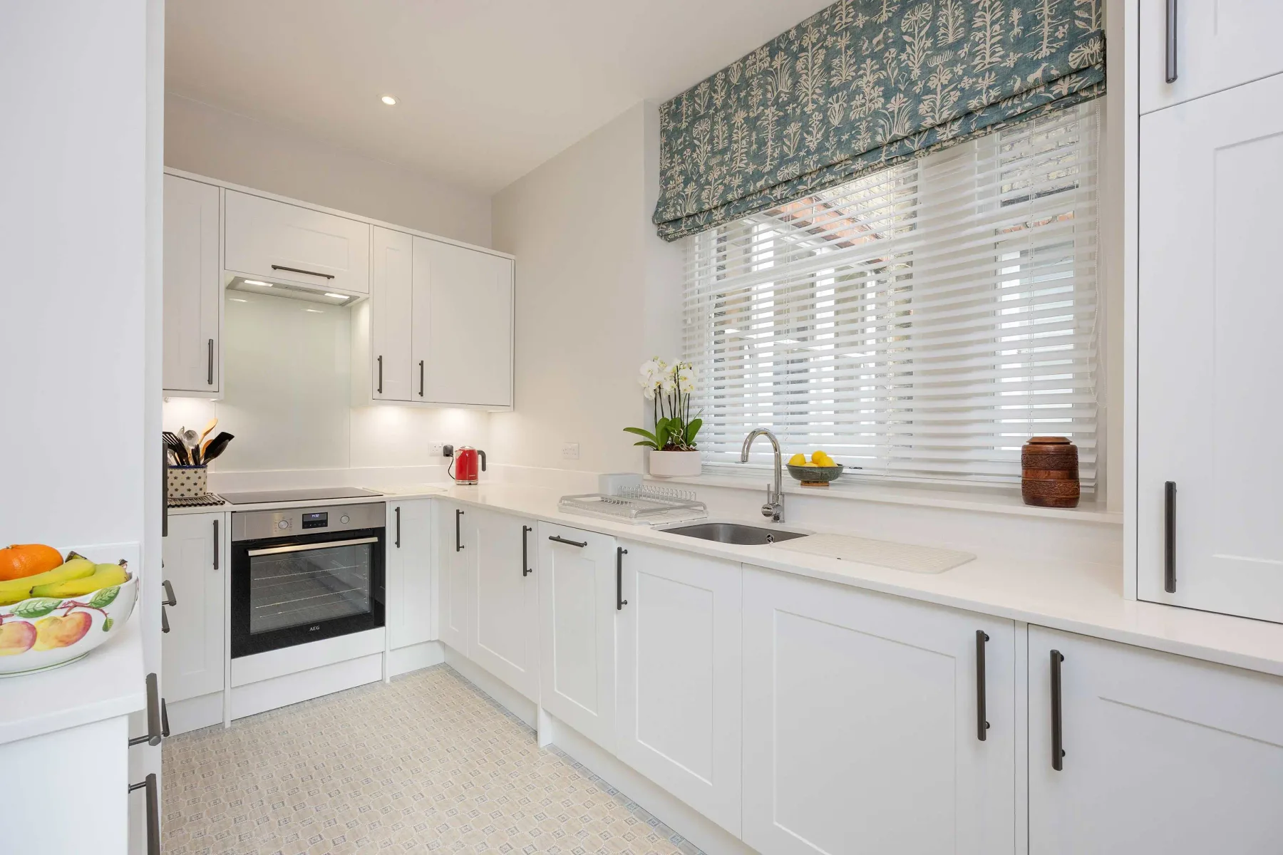 A bright white kitchen with a long counter, sink, and window blinds.