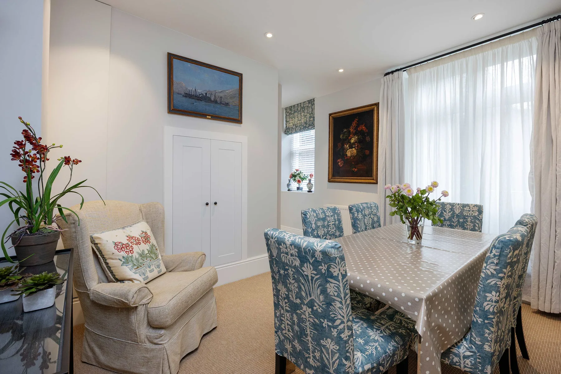 A bright dining area with a patterned tablecloth and upholstered seating beside a sitting area.