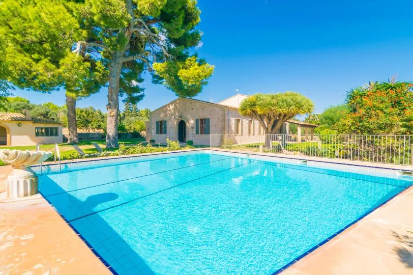 A swimming pool sits beside a stone house and leafy trees under a clear blue sky.