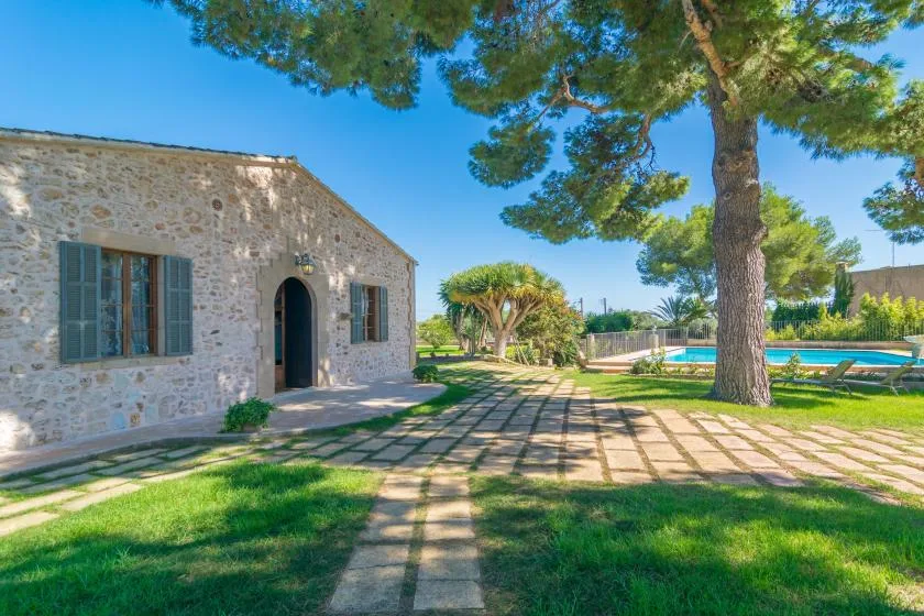A stone building opens onto a paved garden path beneath a large tree.