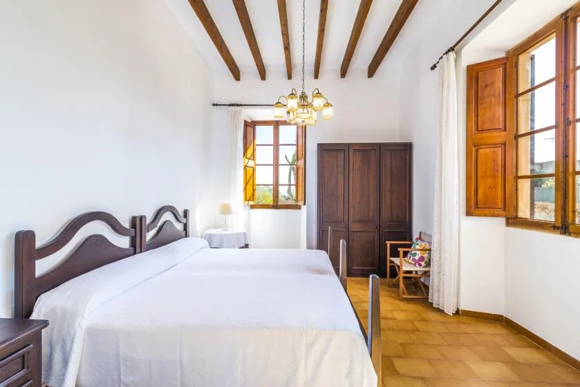 A bright bedroom with a white bed, dark wardrobe, and wooden ceiling beams.