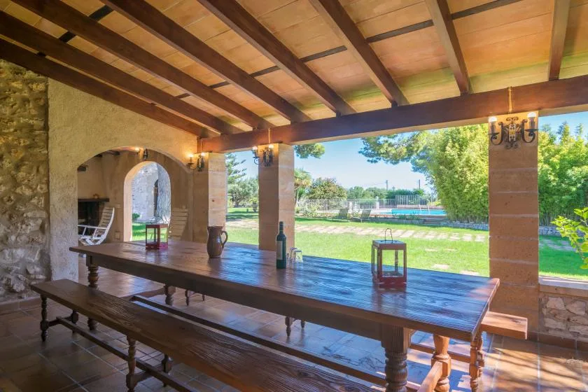 A covered outdoor dining area with a long wooden table overlooking a pool and greenery.