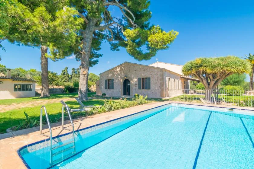 A swimming pool sits beside a stone house, with trees and greenery around it.