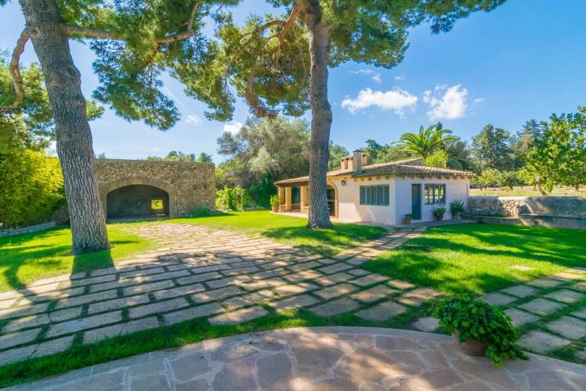 A low house sits beside a paved garden courtyard beneath tall trees.