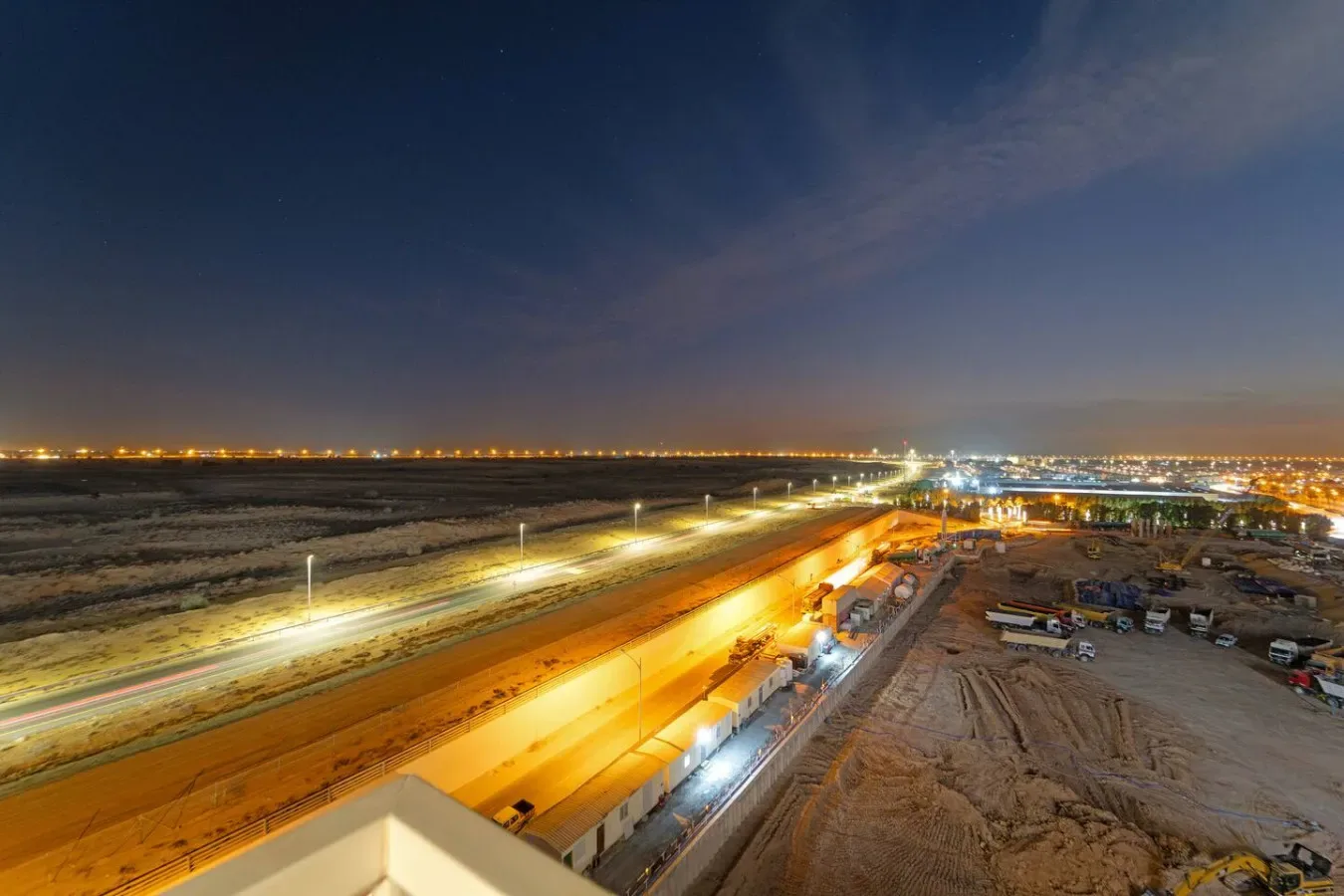 A nighttime construction site beside a brightly lit roadway stretches toward distant city lights.