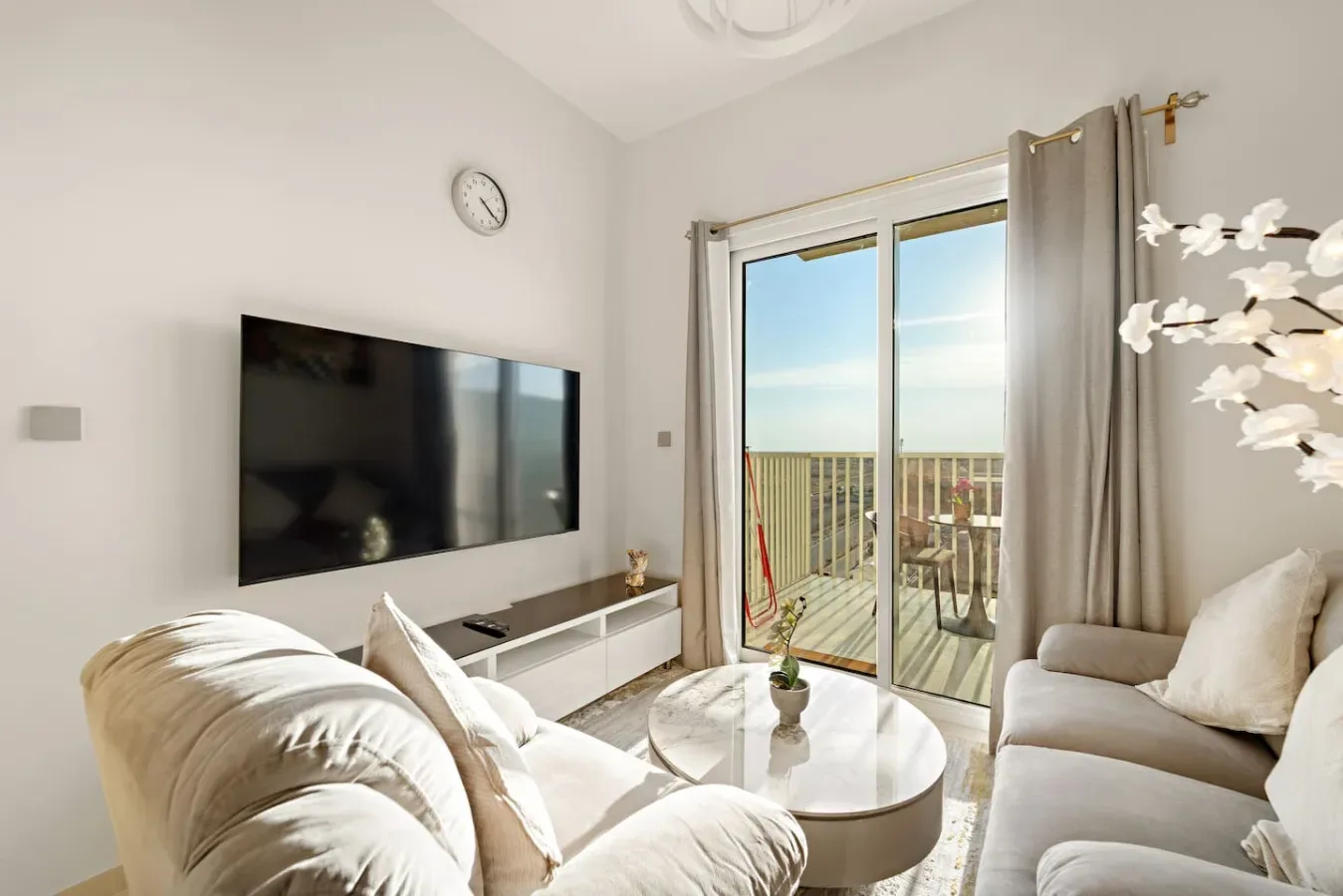 A bright living room with seating, a wall-mounted television, and glass doors opening onto a balcony.