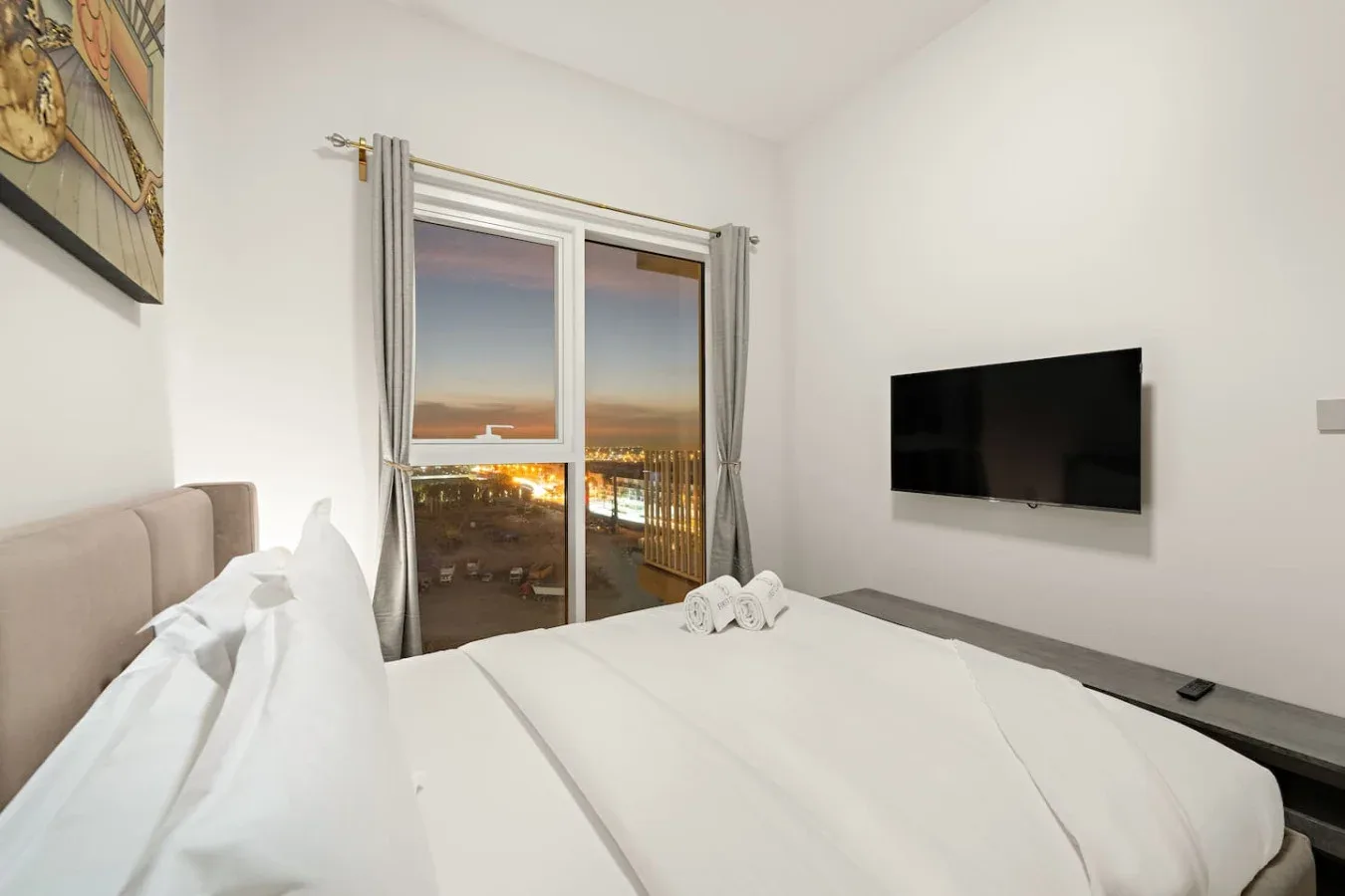 A bright bedroom with a neatly made bed and a window overlooking city lights.
