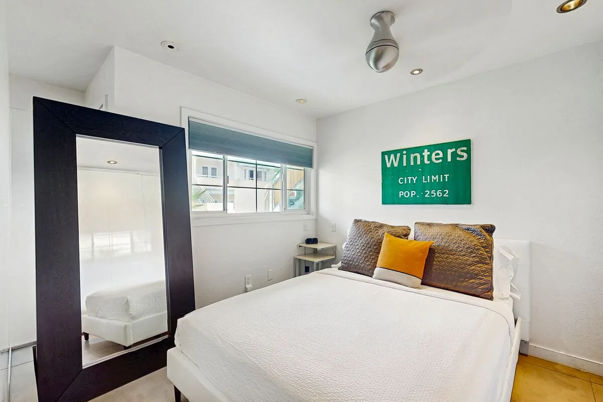 A bright bedroom with a bed, an orange accent pillow, and a window.