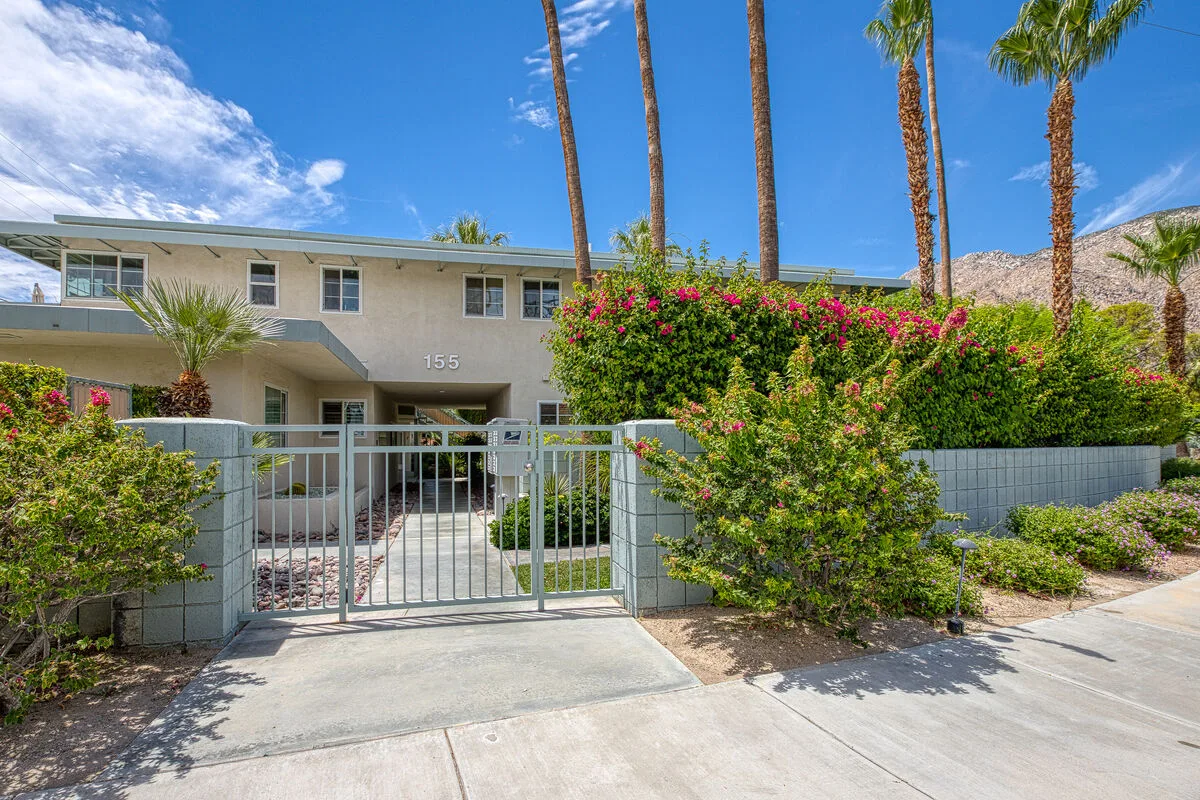 A gated building entrance is surrounded by flowering shrubs and palm trees.