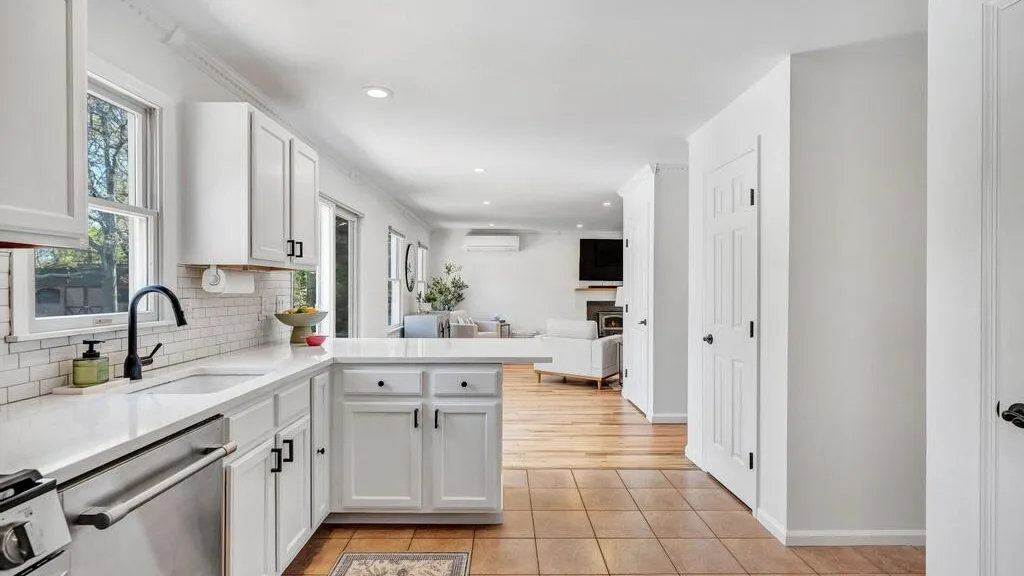 A bright kitchen opens into a living area, with white cabinets and tiled flooring.