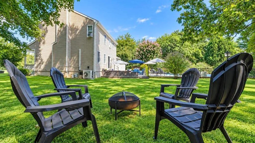 A grassy outdoor seating area with chairs arranged around a fire pit beside a house.