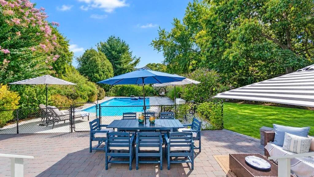 An outdoor poolside patio has a dining table beneath a blue umbrella.